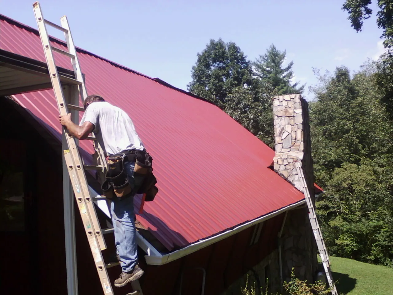 Roofer on a ladder beside a red metal roof and stone chimney — Soffit & Fascia Repair by Roofing Brothers in Wake Forest